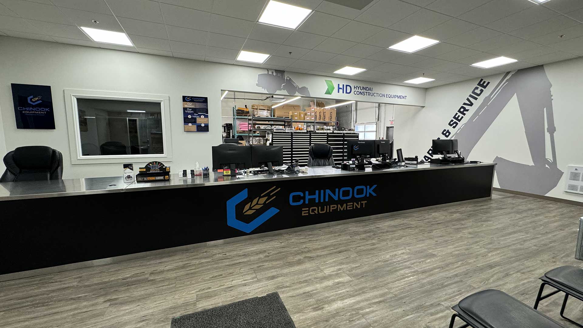 Reception area inside Chinook Equipment showing a long black service counter emblazoned with the Chinook Equipment logo, several computer workstations behind it, a parts storage area visible through a service window labeled “HD Hyundai Construction Equipment,” and a large wall graphic of an excavator arm under bright overhead lighting.